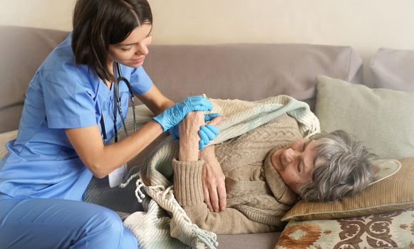 A young nurse is caring for an elderly 80-year-old woman at home, she asks about the well-being of a pensioner who lies and rests in bed. Happy retired woman and trust between doctor and patient.