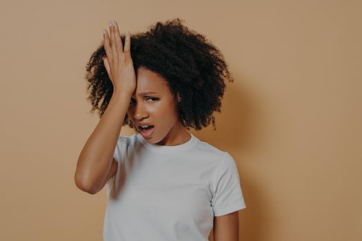 Studio shot of young stressful african woman with hand on forehead suffering from strong headache or head migraine, looking at camera with unpleased face expresion, isolated on sand color background