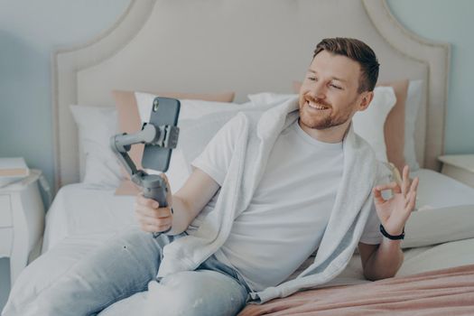 Attractive bearded man in casual clothes lying on bed while chatting online with friends on smartphone, smiling and showing with his hand okey sign, holding cellphone with gimbal stabilizer handheld