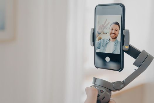 Positive man blogger holding gimbal stabilizer with smartphone and trying to make selfie pictures and live video while spending time at home in living room. Vlog and video blogging concept