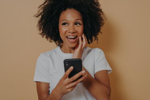 Young smiling happy Afro American woman using cellphone, overjoyed to see, to hear good news, isolated on beige background with copy space for advertising. Positive women emotions and body language