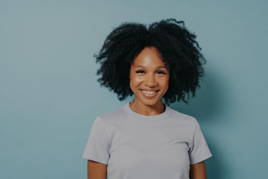 Beautiful smiling african american woman with curly black hair and beaming smile, showing white teeth, wearing casual t-shirt, standing over blue studio wall with copy space, happy to spend day off
