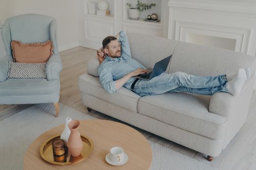 Young handsome successful businessman working remotely at home, compiling data with his notebook laptop while lying on couch with head resting on hand in lightly style living room