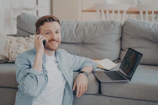 Young male company employee working remotely from home, calling his boss to tell him good news about projects progress, resting hand on couch with laptop and data while sitting on carpet