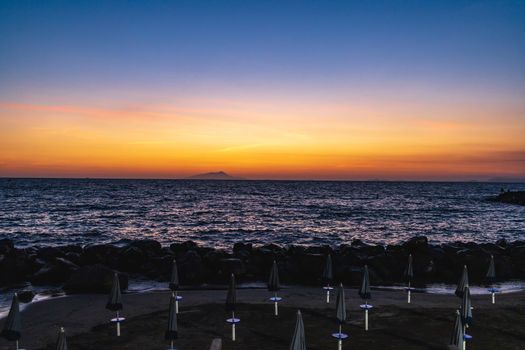 Beautiful sunset over the sea and small beach full of blue and white umbrellas next to giant rocks