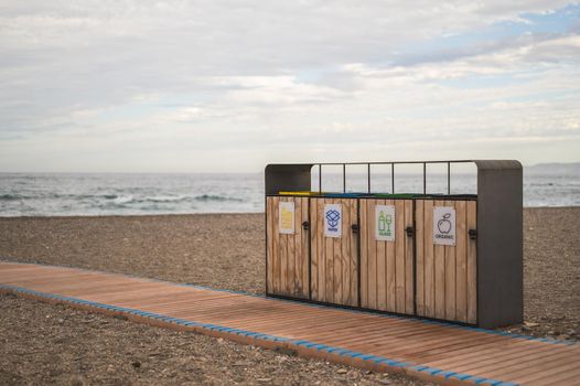 recycling containers in front of a wooden path on the beach with the sea behind and cloudy sky. concept of conservation of the environment