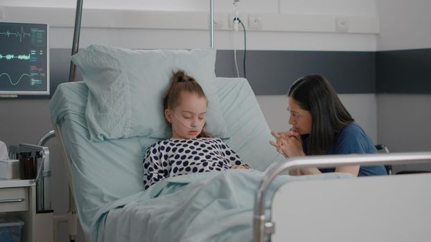 Worried mother sitting beside little daughter praying while sleeping after illness surgery waiting for medical expertise in hospital ward. Sick child wearing nasal tube during medicine treatment