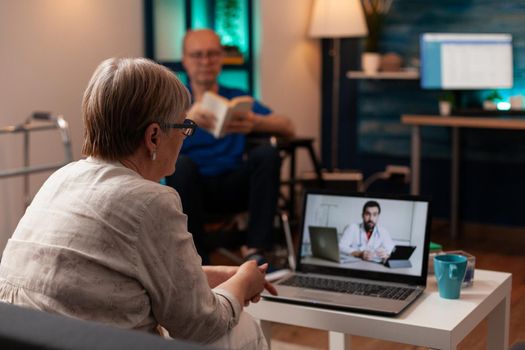 Sick older patient using telemedicine on video call conference while sitting on couch at home. Senior woman discussing healthcare issues with doctor on laptop for remote examination