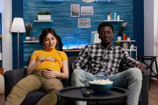 POV of interracial couple expecting child watching television at home. Multi ethnic partners with pregnancy smiling and looking at camera while caucasian pregnant woman touching belly