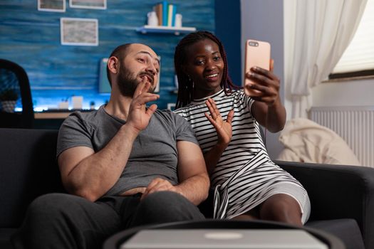Interracial couple waving at camera talking on video call conference. Mixed race people holding modern smartphone with online remote communicatin on internet connection at home.