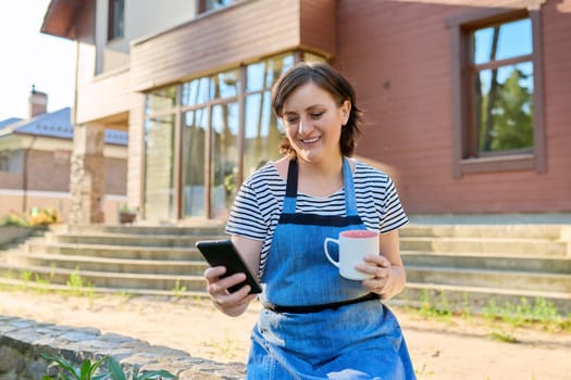 Portrait of happy middle aged woman relaxing outdoors in backyard. Female in garden apron with mug of tea smartphone in her hands, looks at phone screen, spring garden background. 40s people lifestyle