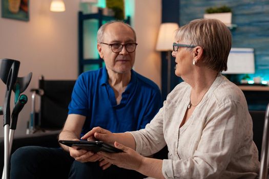 Retired couple using digital tablet and chatting in living room. Senior man and woman sitting together with modern gadget while having crutches and walk frame on couch. Old people with device