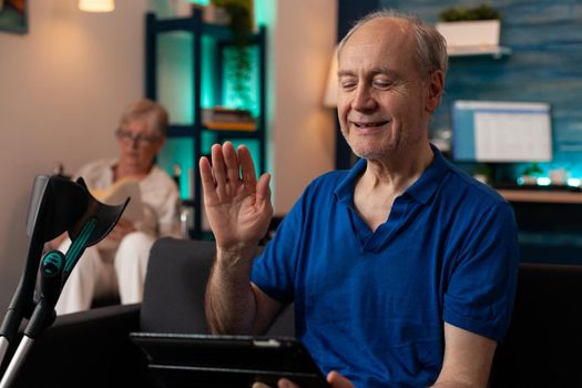 Retired adult waving at tablet with video call conference on sofa with crutches. Old man using online remote communication while elder disabled woman sitting in wheelchair reading