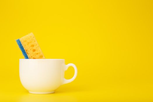Creative, minimal dishwashing concept. Simple composition with yellow cleaning kitchen sponge in white ceramic cup on yellow background with copy space. 
