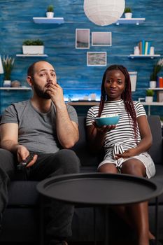 Interracial couple sitting on couch watching television at home. African american woman holding bowl with popcorn while caucasian man switching channels with TV remote control.
