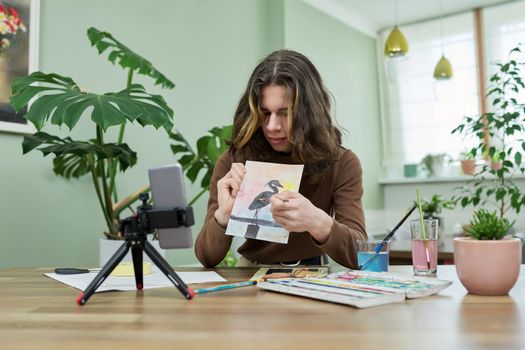 Guy teenager student drawing online using smartphone. Teenage male with watercolors, brushes, sketch, sitting at home, showing picture on phone. Video lesson, virtual meeting with teacher, creativity