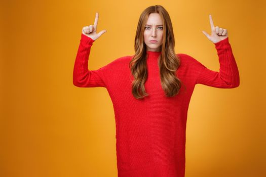 Intense disappointed and distressed attractive young 20s redhead female in red warm sweater frowning upset with dismay raising hands pointing up dissatisfied against orange background.