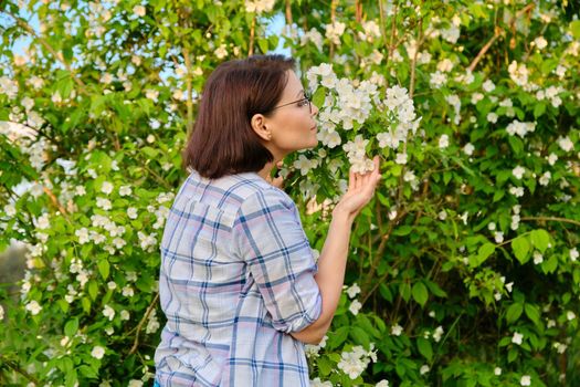 Portrait of smiling middle-aged woman near flowering jasmine bush. Beautiful female 40s of age enjoying scent of flowers in spring garden. Nature, springtime, beauty, people of mature age concept