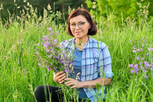 Portrait of middle-aged woman with bouquet of wildflowers in nature. Female 40s age with bells in hands looking at camera, green spring summer grass meadow background. Beauty, nature, people of mature age