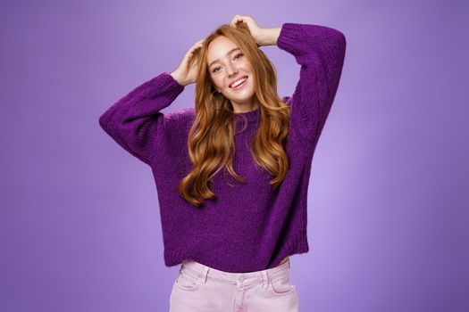 Carefree joyful and cool stylish redhead female student in purple warm sweater enjoying holidays stretching, holding hands near head, tilting and smiling happily relaxing against violet background.