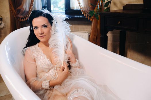the bride, dressed in a boudoir transparent dress and underwear, lies in a vintage bathroom with a white feather in her hands, The morning of the bride.