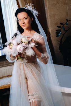 the bride, dressed in a boudoir transparent dress and underwear, sits near a vintage bath with a feather in her hands, The morning of the bride.