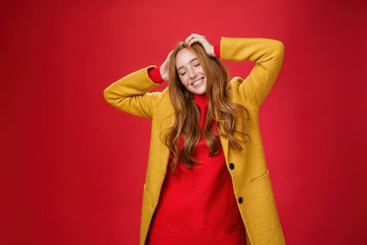 Carefree happy ginger girl carrying away with music and good vibes holding hands on head close eyes and smiling delighted and relaxed having fun, dancing against red background in yellow coat.