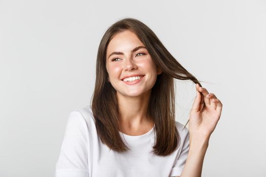 Portrait of flirty beautiful girl playing with hair and smiling at camera coquettish, standing white background.