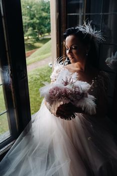 A bride in a wedding dress and a bouquet sits at an open old window and looks.