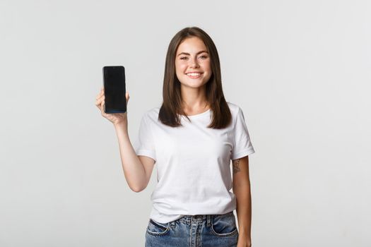 Attractive smiling young woman looking pleased, showing smartphone screen.