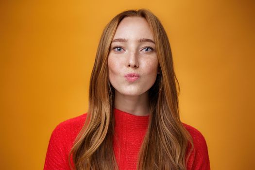 Close-up shot of flirty good-looking sensual redhead female in red knitted clothes folding lips in kiss and gazing at camera as giving mwah feleing romantic and pretty against orange background. Body language, fashion and people concept