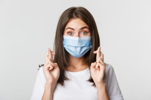 Covid-19, health and social distancing concept. Close-up of hopeful smiling girl in medical mask, cross fingers good luck.