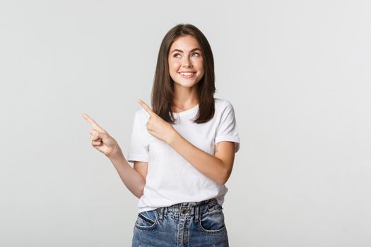 Dreamy beautiful girl smiling, pointing fingers upper left corner and looking pleased at logo.