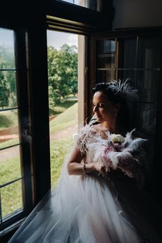 A bride in a wedding dress and a bouquet sits at an open old window and looks.
