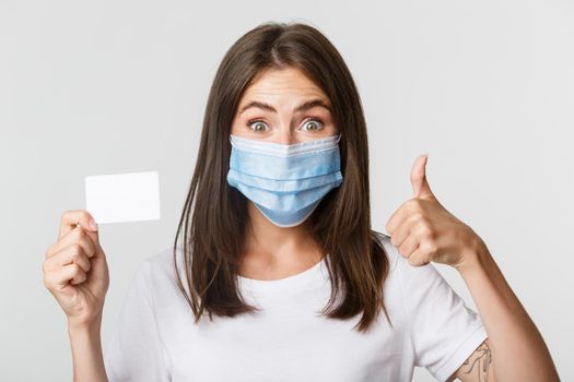 Covid-19, health and social distancing concept. Close-up of pretty brunette girl in medical mask, showing credit card and thumbs-up in approval.