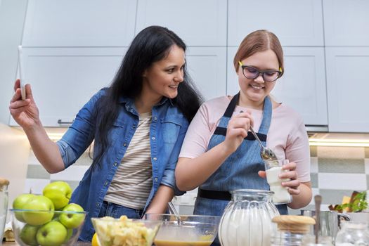 Mom and teen daughter preparing apple pie together, at home in kitchen. Woman with digital tablet, recipe, video call, technology in daily life. Family, parent teenager relationship, lifestyle