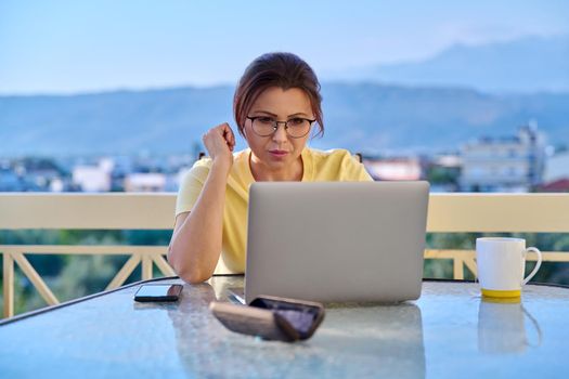 Serious middle aged woman with laptop at table on outdoor balcony. Concentrated 40s mature business woman working remotely. Freelance, technology, work from home office, online training, blog, vlog