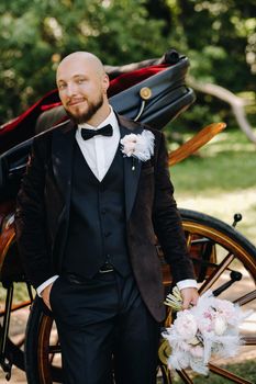 The groom with a bouquet in his hands stands near the carriage in nature in retro style.
