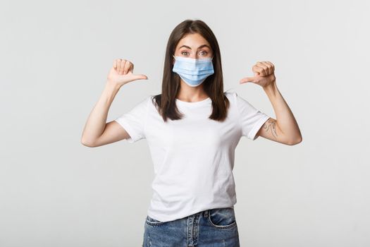 Covid-19, health and social distancing concept. Attractive smiling woman in medical mask pointing at herself, white background.