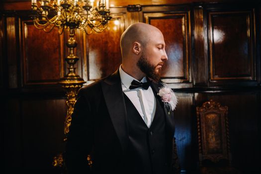 Portrait of the groom in a suit and bow tie, standing in the interior.