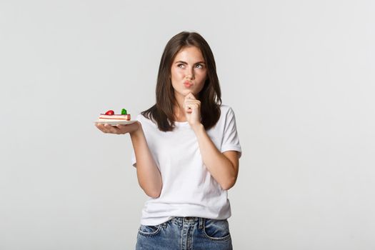 Thoughtful smiling pretty girl pondering while holding piece of cake, white background.
