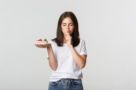 Thoughtful smiling pretty girl pondering while holding piece of cake, white background.