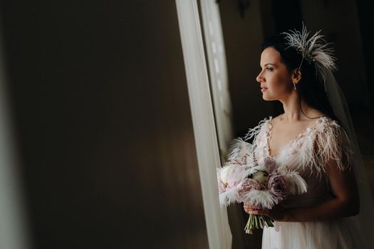 The bride in a wedding dress and with a bouquet stands at the old window and looks.