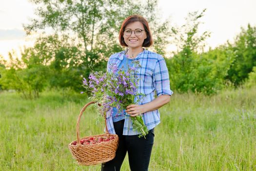 Portrait of middle-aged woman with bouquet of wildflowers and basket of strawberries in nature. Female 40s age looking at camera, green spring summer grass meadow background