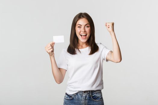 Rejoicing beautiful girl showing credit card and triumphing, fist pump happy, white background.