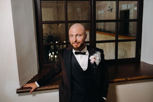 Portrait of the groom in a suit and bow tie, standing in the interior.
