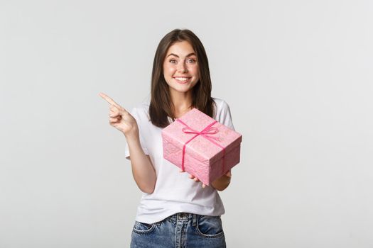 Happy smiling brunette girl holding birthday gift and pointing finger left at logo.