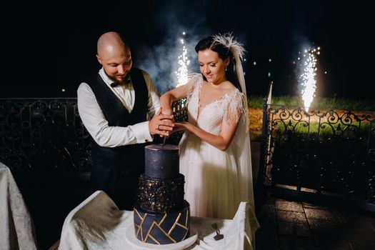 Stylish bride and groom cut the wedding cake in the evening on the street.