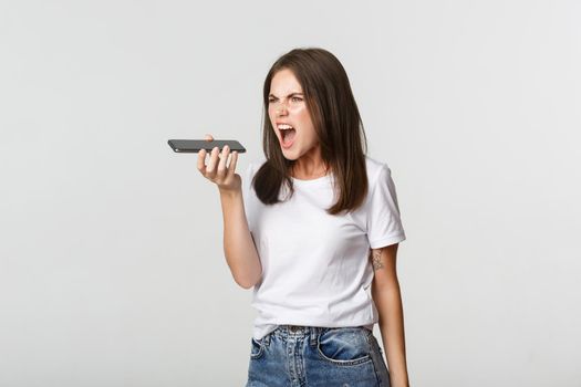 Angry brunette girl shouting mad while recording voice message, being outraged, white background.