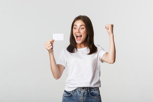 Joyful good-looking girl rejoicing and looking at credit card, fist pump while triumphing, white background.